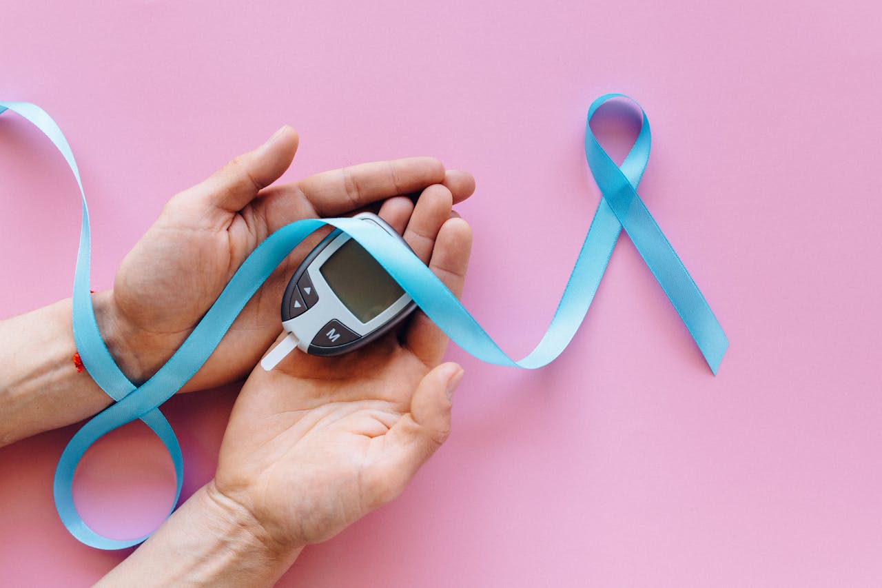 A close-up image of hands holding a glucometer with a blue awareness ribbon on a pink surface, symbolizing diabetes awareness.